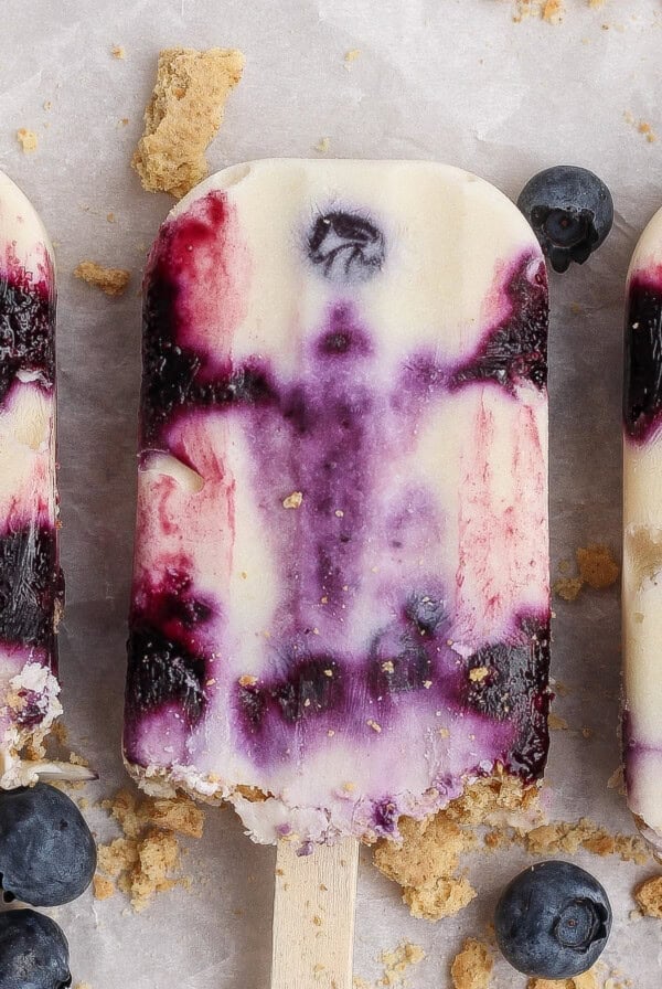 Three creamy blueberry popsicles with purple swirls are arranged on a light surface, surrounded by scattered blueberries and crumbled cookie pieces.