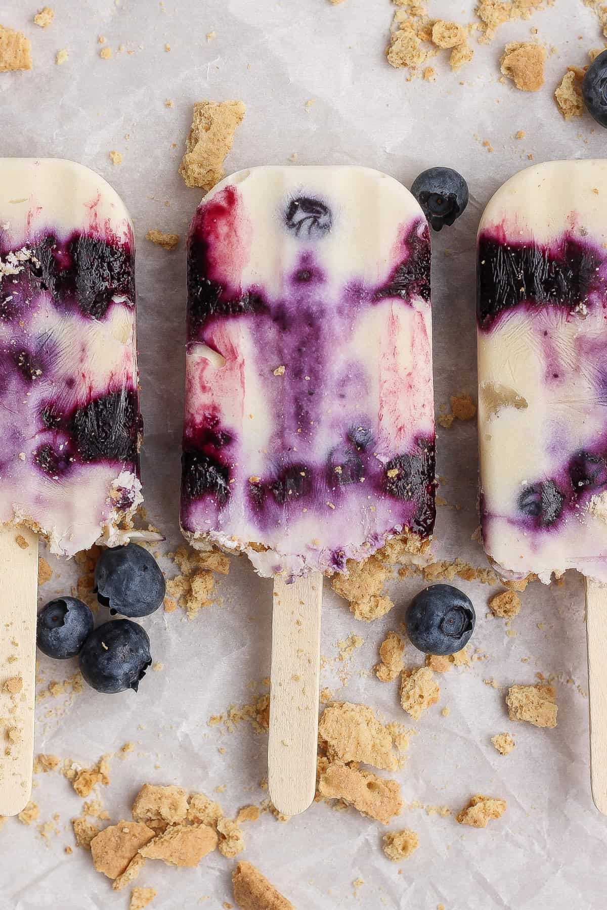 Three blueberry cheesecake popsicles with visible blueberry swirls are arranged on a surface scattered with fresh blueberries and crumbled graham crackers.