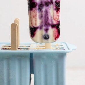 A hand holds a popsicle with purple, white, and red swirls above a blue silicone popsicle mold containing more popsicles.