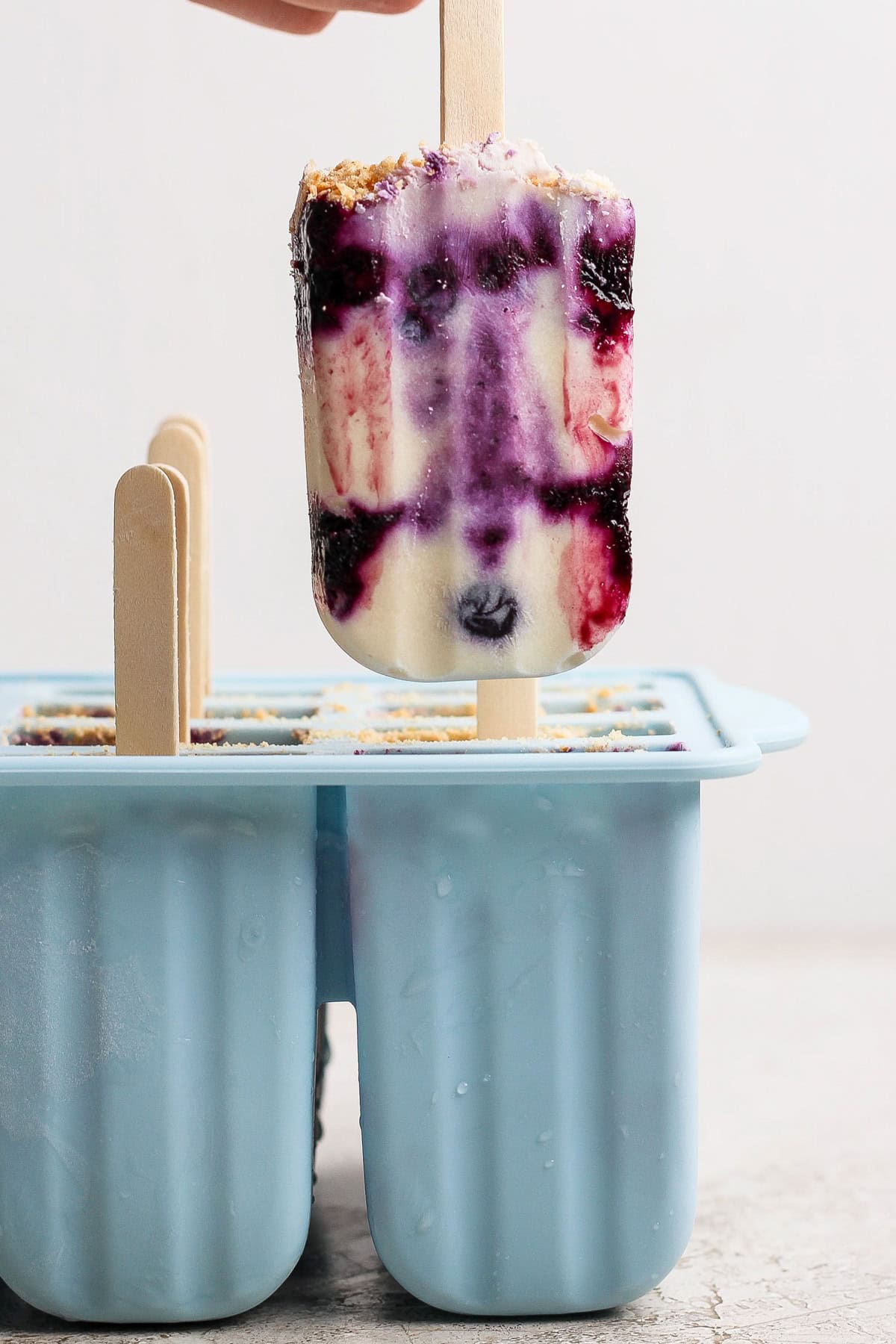 A hand holds a popsicle with purple, white, and red swirls above a blue silicone popsicle mold containing more popsicles.