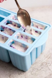A spoon fills a blue silicone mold tray with a white and purple mixture on a light-colored surface.