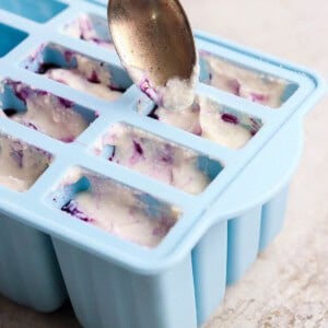 A spoon fills a blue silicone mold tray with a white and purple mixture on a light-colored surface.