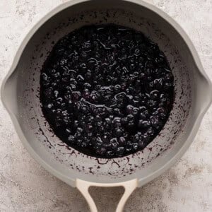 A beige saucepan filled with cooked blueberries in a thick purple sauce on a light textured surface.