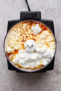 A food processor bowl filled with cottage cheese, eggs, flour, and vanilla extract, viewed from above.