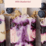 A cottage cheese popsicle with visible swirls of blueberry and crumbs, surrounded by blueberries and broken graham crackers on a light surface.