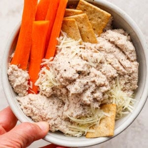 A bowl containing carrot sticks, crackers, shredded cheese, and a creamy dip, held in one hand.