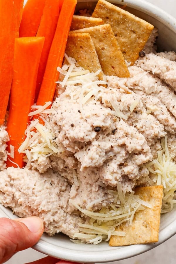 A bowl containing carrot sticks, crackers, shredded cheese, and a creamy dip, held in one hand.