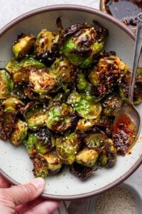A bowl of roasted Brussels sprouts topped with sesame seeds and chili oil, with a spoon on the side.