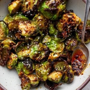 A bowl of roasted Brussels sprouts topped with sesame seeds and chili oil, with a spoon on the side.