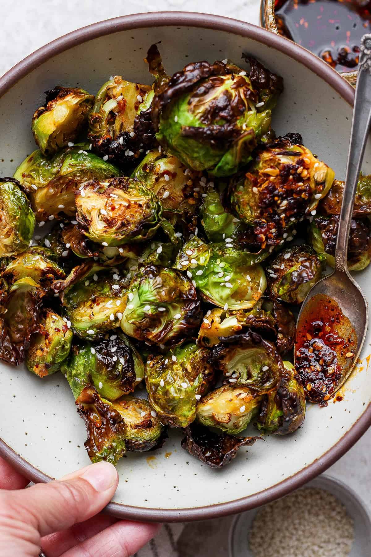A bowl of roasted Brussels sprouts topped with sesame seeds and chili oil, with a spoon on the side.
