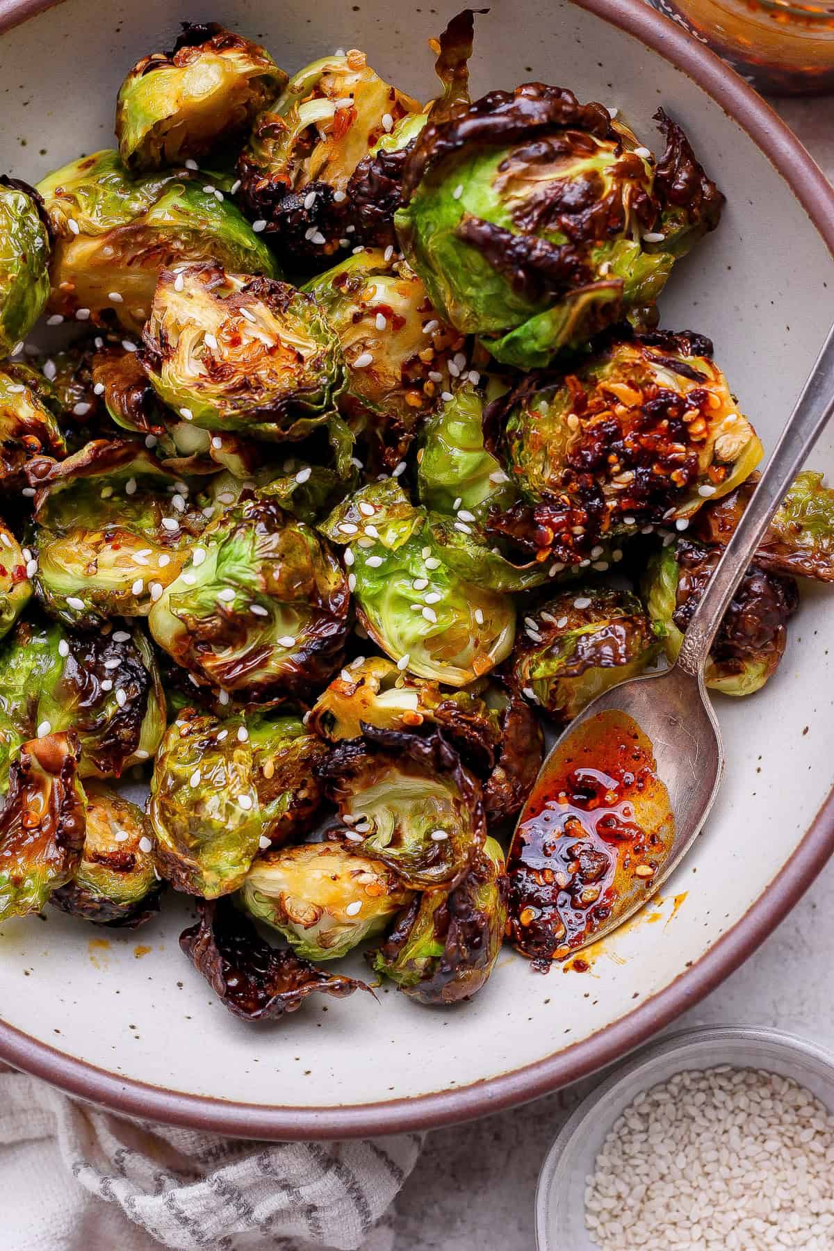 A bowl of roasted Brussels sprouts garnished with sesame seeds, served with a spoon and chili oil.