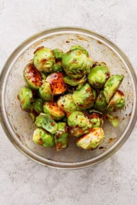 A glass bowl filled with halved Brussels sprouts coated in a dark seasoning or marinade, placed on a light textured surface.