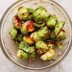 A glass bowl filled with halved Brussels sprouts coated in a dark seasoning or marinade, placed on a light textured surface.