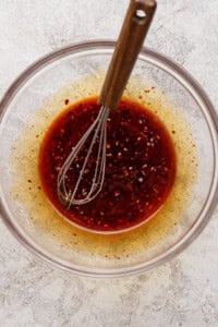 A glass bowl containing a reddish-brown liquid with visible spices and a metal whisk with a wooden handle resting inside.