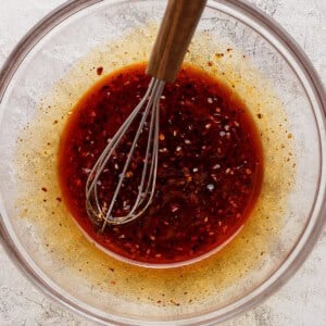 A glass bowl containing a reddish-brown liquid with visible spices and a metal whisk with a wooden handle resting inside.