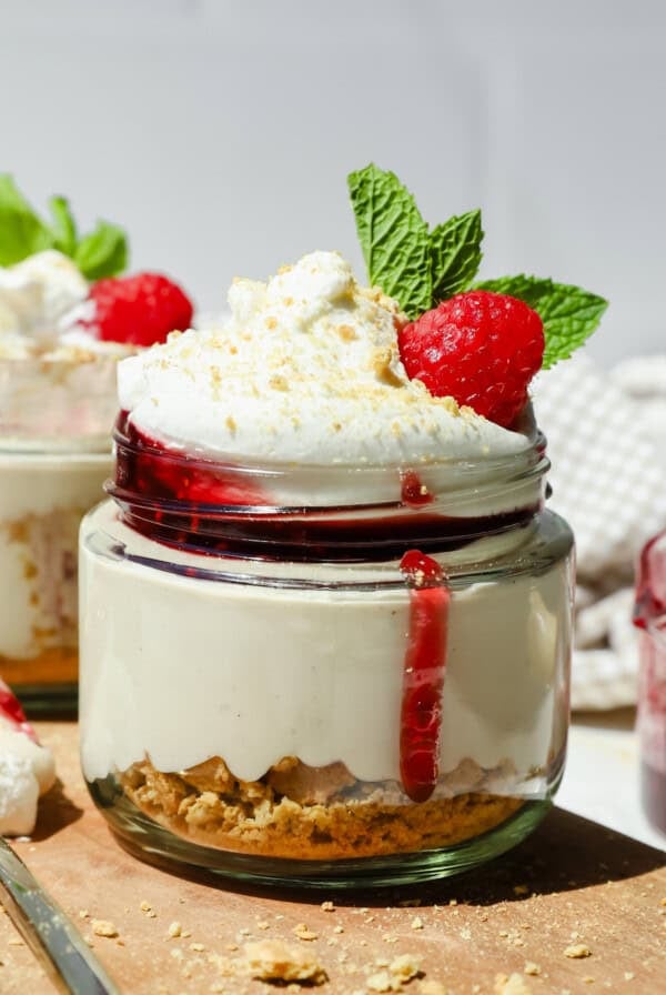 A small glass jar of cheesecake topped with whipped cream, raspberry sauce, a fresh raspberry, and mint leaves, with a layer of crushed biscuits at the bottom.
