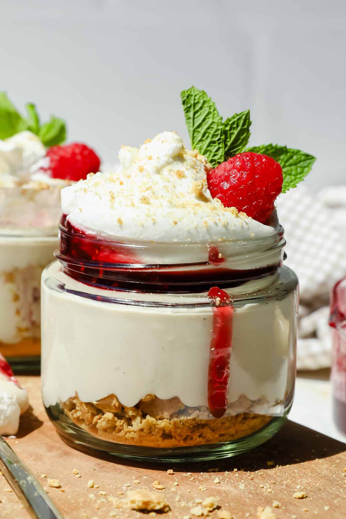 A small glass jar of cheesecake topped with whipped cream, raspberry sauce, a fresh raspberry, and mint leaves, with a layer of crushed biscuits at the bottom.