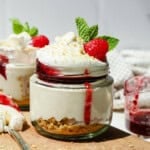 A dessert jar with layered cheesecake, graham cracker crust, whipped cream, raspberry sauce, a raspberry, and a mint leaf on top.