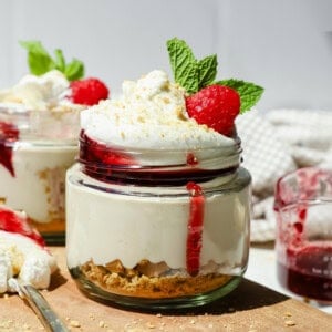 A dessert jar with layered cheesecake, graham cracker crust, whipped cream, raspberry sauce, a raspberry, and a mint leaf on top.