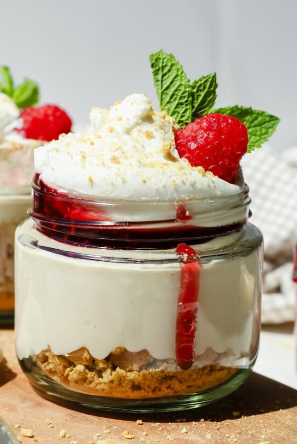 A dessert jar with layered cheesecake, graham cracker crust, whipped cream, raspberry sauce, a raspberry, and a mint leaf on top.