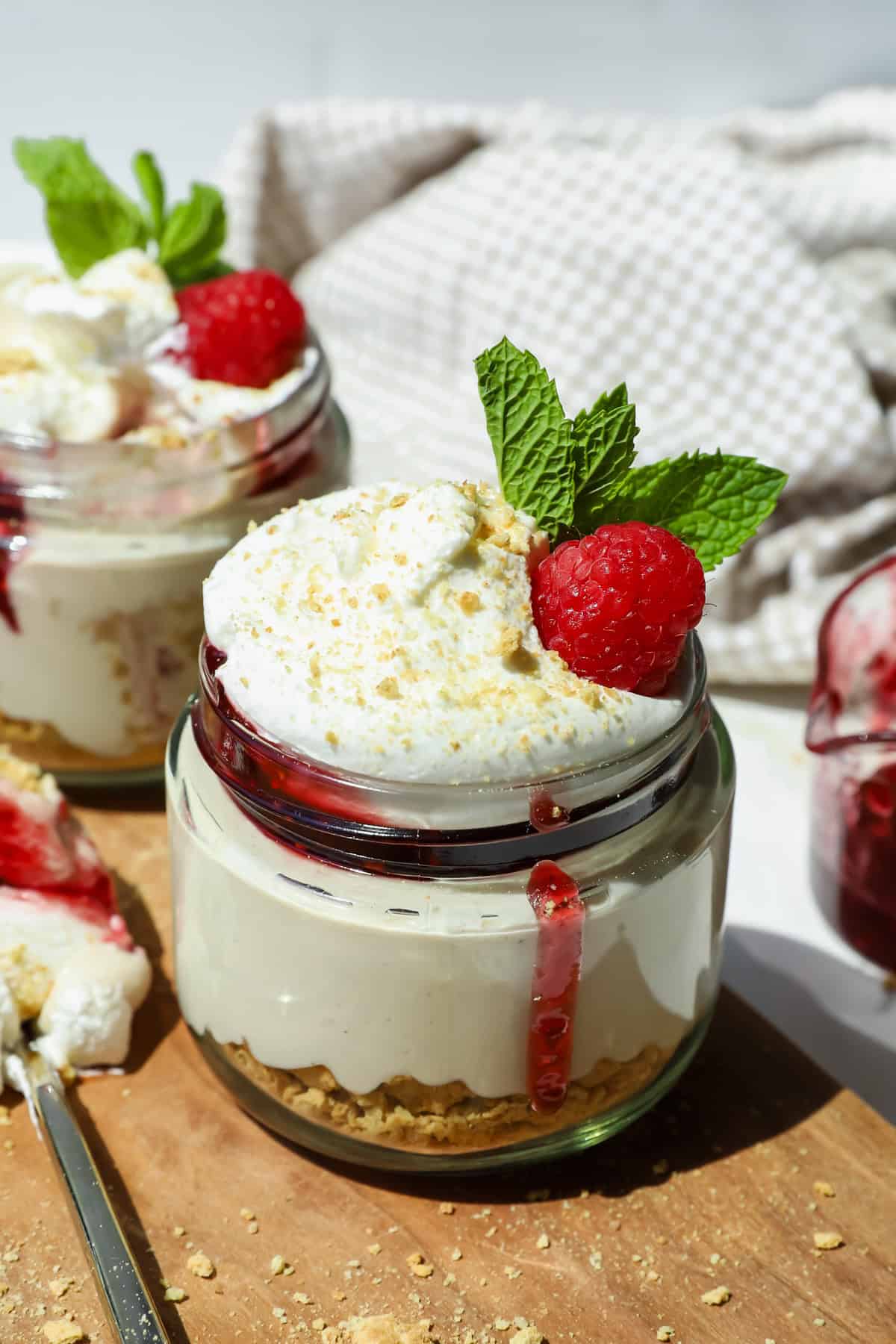 A glass jar filled with layered cheesecake, graham cracker crust, whipped cream, a raspberry, and mint leaves, with berry sauce dripping down the side.