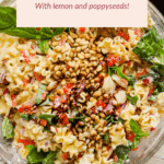 Bowl of cottage cheese pasta salad with lemon, poppyseeds, chopped vegetables, fresh greens, and a topping of pine nuts.