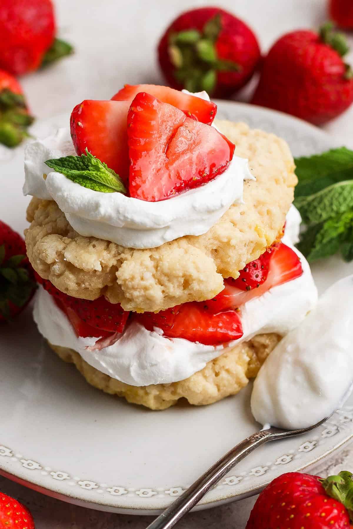 Un postre de pastel de fresa con capas de galletas, crema batida y fresas frescas, adornadas con una hoja de menta, en un plato blanco con una cuchara y fresas enteras.