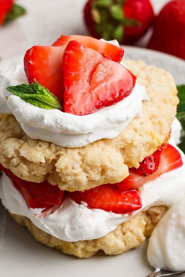 A strawberry shortcake dessert with layers of biscuits, whipped cream, and sliced strawberries, garnished with mint leaves on a white plate.