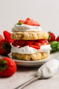 A strawberry shortcake dessert with layers of biscuits, whipped cream, and sliced strawberries on a plate, surrounded by whole strawberries and mint leaves.