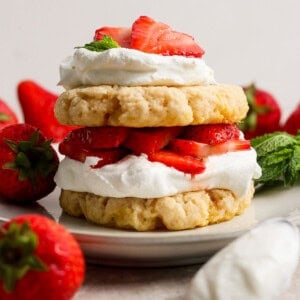 A strawberry shortcake dessert with layers of biscuits, whipped cream, and sliced strawberries on a plate, surrounded by whole strawberries and mint leaves.