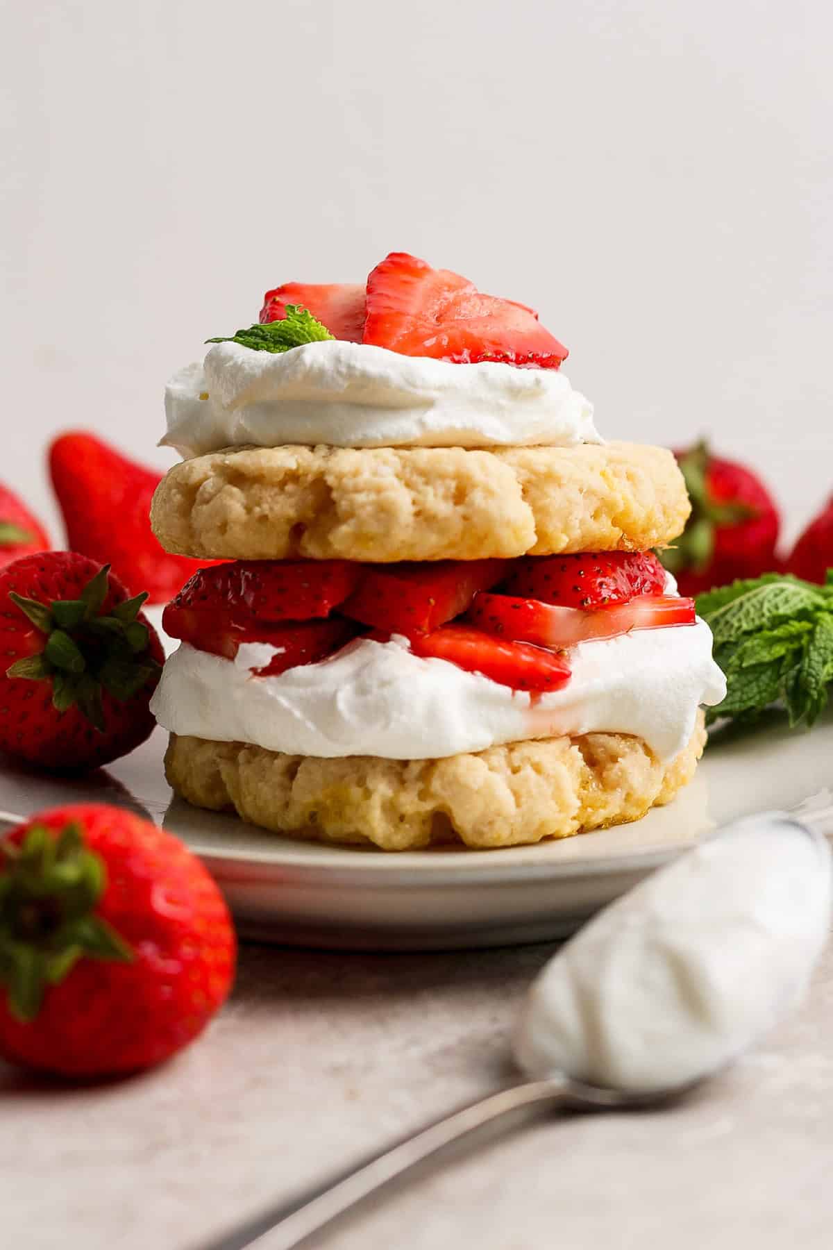 Un postre de pastel de fresa con capas de galletas, crema batida y fresas en rodajas en un plato, rodeado de fresas enteras y hojas de menta.