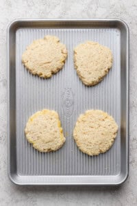 Four unbaked biscuit dough rounds are spaced evenly on a metal baking sheet placed on a light-colored surface.