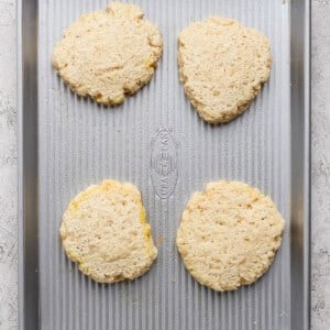 Four unbaked biscuit dough rounds are spaced evenly on a metal baking sheet placed on a light-colored surface.