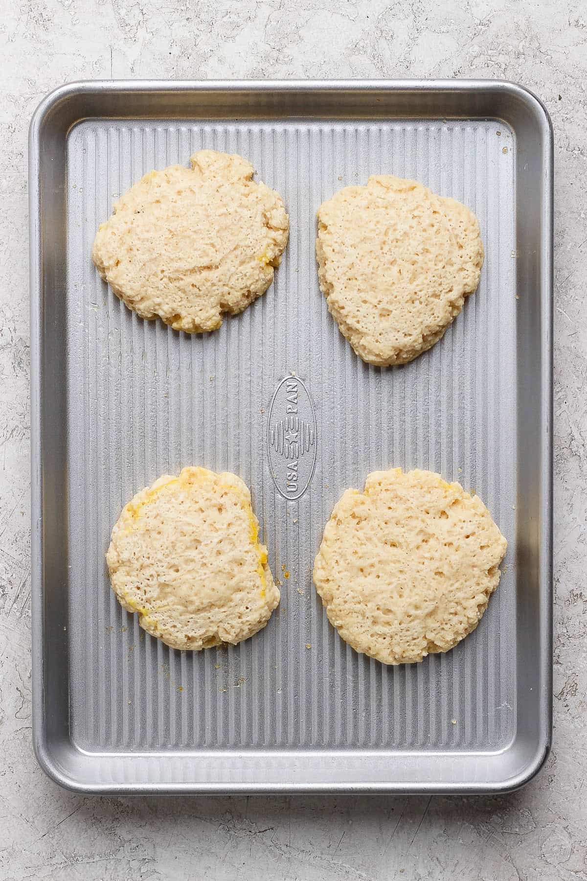 Cuatro rondas de masa de galletas sin hornear están espaciadas uniformemente sobre una bandeja para hornear metal colocada sobre una superficie de color claro.