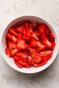 A white bowl filled with sliced fresh strawberries sits on a light textured surface.