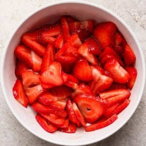 A white bowl filled with sliced fresh strawberries sits on a light textured surface.