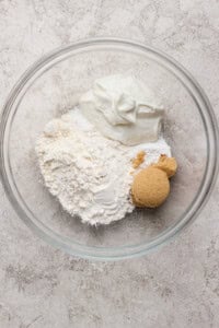 A glass bowl containing flour, brown sugar, and a scoop of white yogurt on a light textured surface.