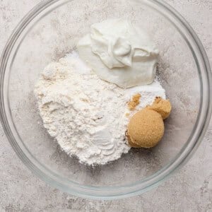 A glass bowl containing flour, brown sugar, and a scoop of white yogurt on a light textured surface.