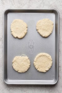 Four unbaked biscuit dough rounds evenly spaced on a silver baking sheet, placed on a light-colored surface.