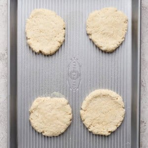Four unbaked biscuit dough rounds evenly spaced on a silver baking sheet, placed on a light-colored surface.