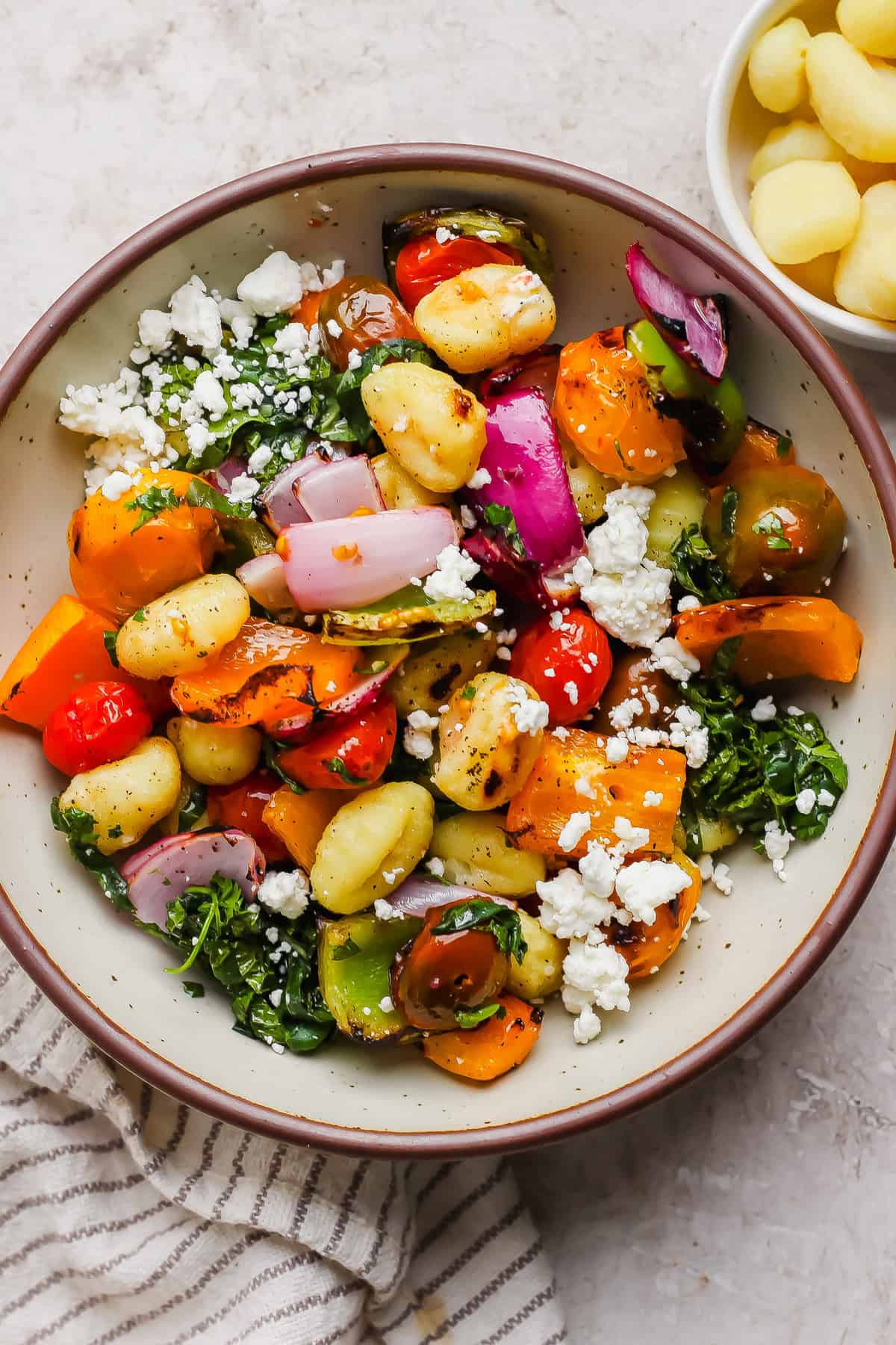 A bowl of gnocchi mixed with roasted vegetables, red onion, tomatoes, leafy greens, and crumbled feta cheese, with a small bowl of gnocchi on the side.