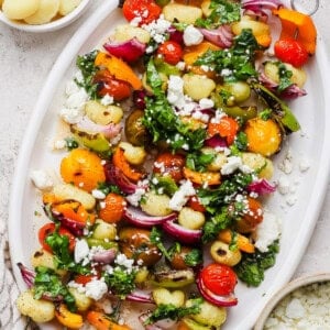 A white platter with gnocchi, roasted cherry tomatoes, bell peppers, red onions, fresh greens, and crumbled feta cheese.