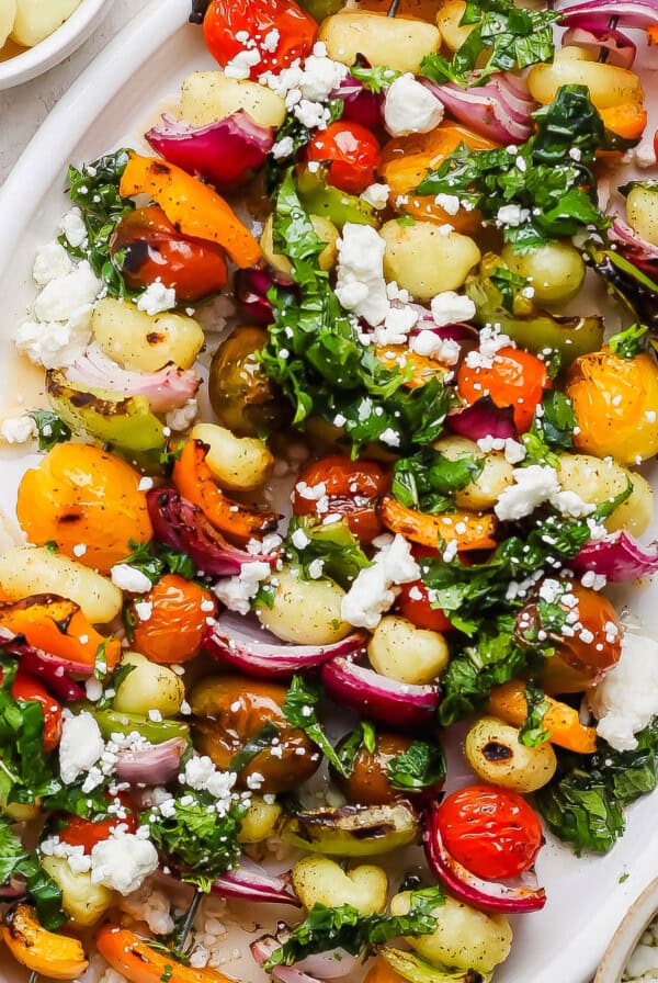 A white platter with gnocchi, roasted cherry tomatoes, bell peppers, red onions, fresh greens, and crumbled feta cheese.