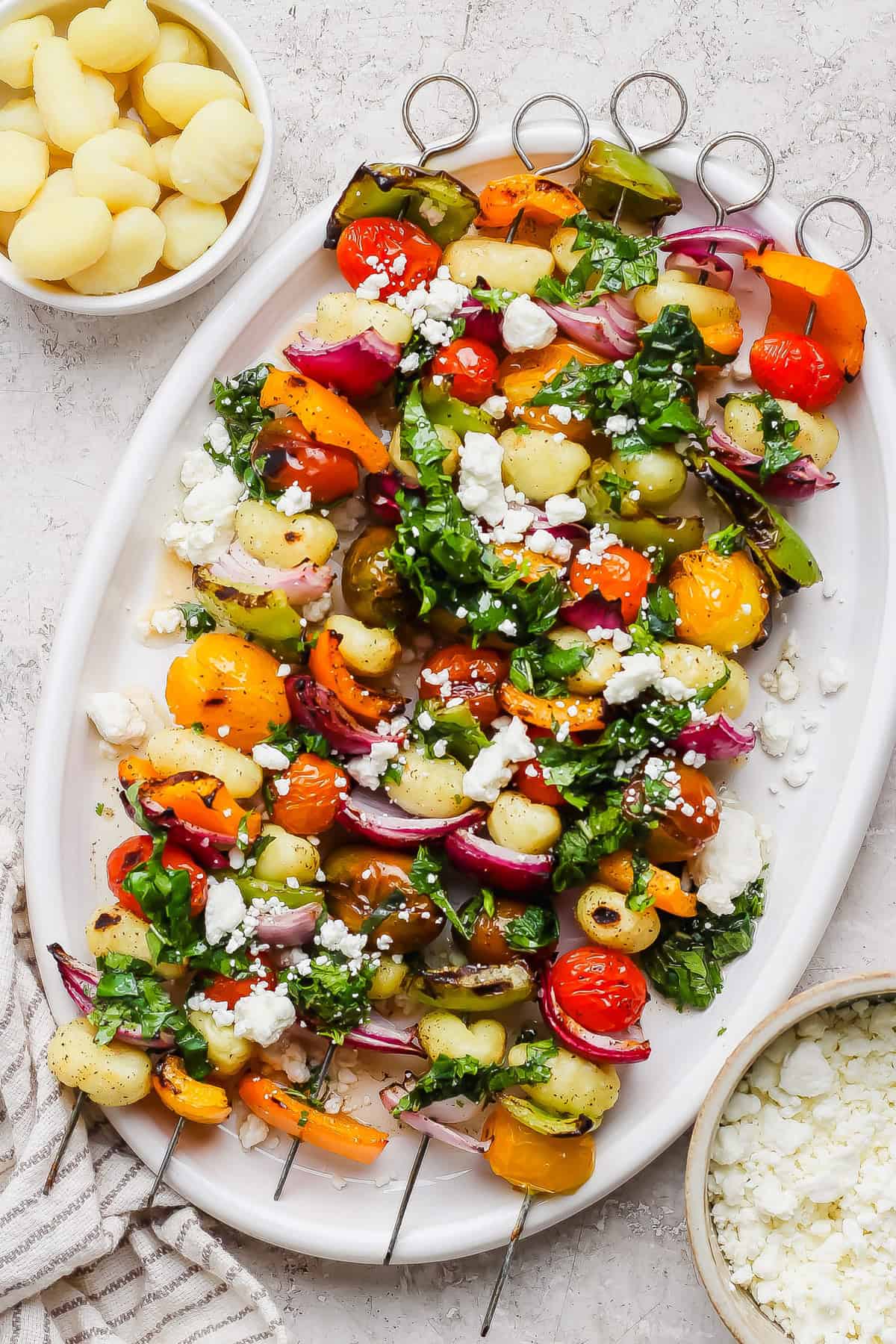 A white platter with vegetable and gnocchi skewers, topped with chopped herbs and crumbled cheese, is pictured next to small bowls of cooked gnocchi and crumbled cheese.