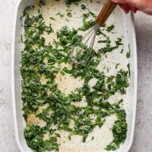 A hand uses a whisk to mix chopped greens and liquid with seasonings in a white rectangular baking dish.