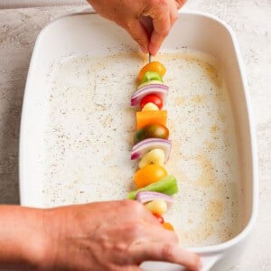 A person places a skewer with vegetables—tomatoes, onions, peppers—into a white baking dish.