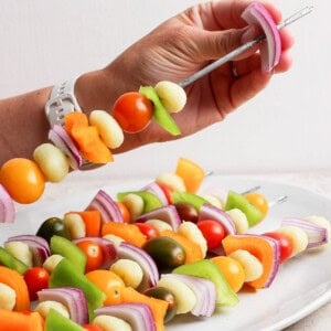 A hand holds a metal skewer with pieces of onion, bell pepper, cherry tomatoes, and other vegetables above a plate of similar vegetable skewers.