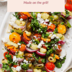 Grilled gnocchi skewers with cherry tomatoes, bell peppers, and red onions are topped with herbs and crumbled cheese on a white platter.
