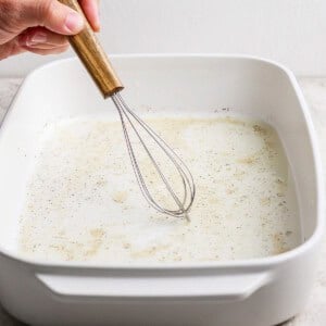 A hand uses a wooden-handled whisk to mix a liquid with spices in a white rectangular baking dish on a light countertop.