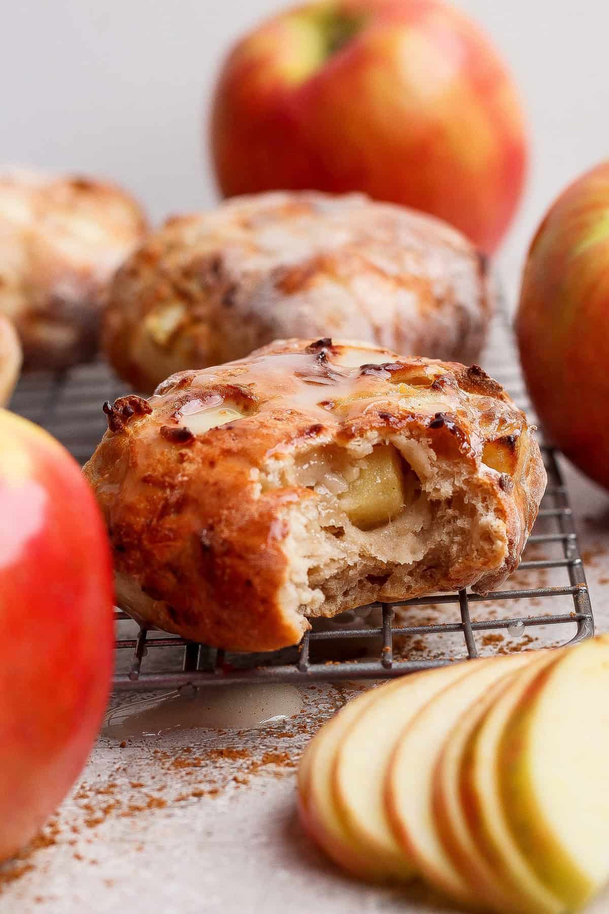 A partially eaten apple scone sits on a cooling rack, surrounded by whole apples and apple slices.